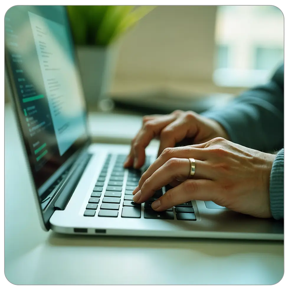 Zwei Hände, die auf der Tastatur eines silbernen Laptops tippen. Der Bildschirm zeigt Programmiercode. Im Hintergrund ist ein unscharf dargestellter grüner Pflanzenkübel und ein Fenster mit natürlichem Licht sichtbar.